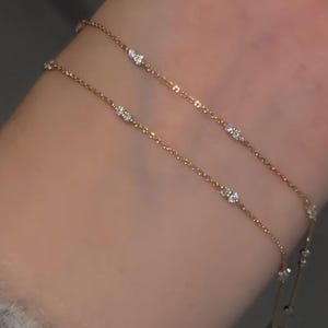 Puede incluir: Una delicada pulsera dorada con pequeñas gemas brillantes. La pulsera presenta dos hilos de fina cadena de oro, cada uno adornado con gemas claras espaciadas uniformemente. La pulsera se usa en una muñeca, mostrando su elegante diseño.