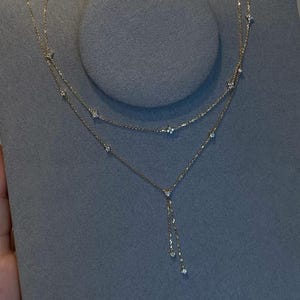 May include: A delicate gold necklace with a layered design. The necklace features small, sparkling gemstones and a dangling chain with three additional gems. The jewelry is displayed on a grey surface.
