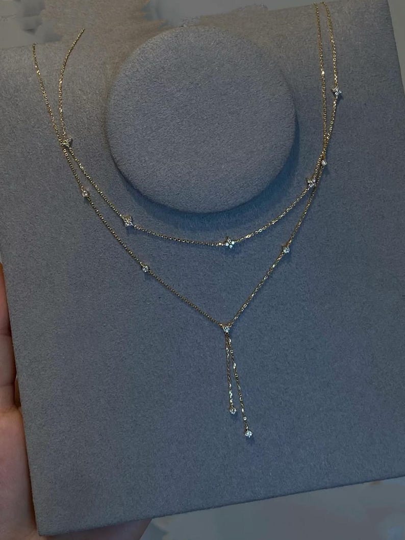 May include: A delicate gold necklace with a layered design. The necklace features small, sparkling gemstones and a dangling chain with three additional gems. The jewelry is displayed on a grey surface.