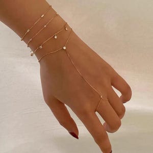 Könnte beinhalten: Eine goldene Handkette bestehend aus drei zarten Armbändern, die mit einem Ring verbunden sind. Der Schmuck besteht aus dünnen Goldketten, die mit kleinen, gleichmäßig verteilten klaren Steinen besetzt sind. Das Design umschließt das Handgelenk und reicht bis zum Mittelfinger.