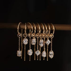 Puede incluir: Un conjunto de pendientes de aro dorados con diversas gemas colgantes en forma de diamante y ovalada. Los pendientes se exhiben en una barra de madera sobre un fondo oscuro, mostrando sus intrincados diseños y detalles brillantes.