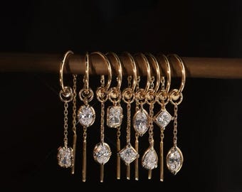 Op de afbeelding: Een verzameling gouden oorringen met verschillende diamant- en ovaalvormige edelstenen. De oorbellen worden getoond op een houten staaf tegen een donkere achtergrond, waarbij hun ingewikkelde ontwerpen en sprankelende details worden benadrukt.