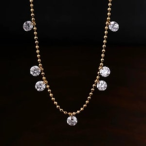 Puede incluir: Un delicado collar de oro con múltiples piedras preciosas redondas y transparentes. El collar presenta una fina cadena de pequeñas cuentas de oro, con piedras preciosas más grandes espaciadas uniformemente a lo largo de la cadena. El fondo es oscuro.
