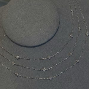 May include: Multiple delicate silver necklaces with small, sparkling flower-shaped pendants are displayed on a grey surface. The necklaces are layered, creating a tiered effect. The chains are fine and the pendants are evenly spaced.