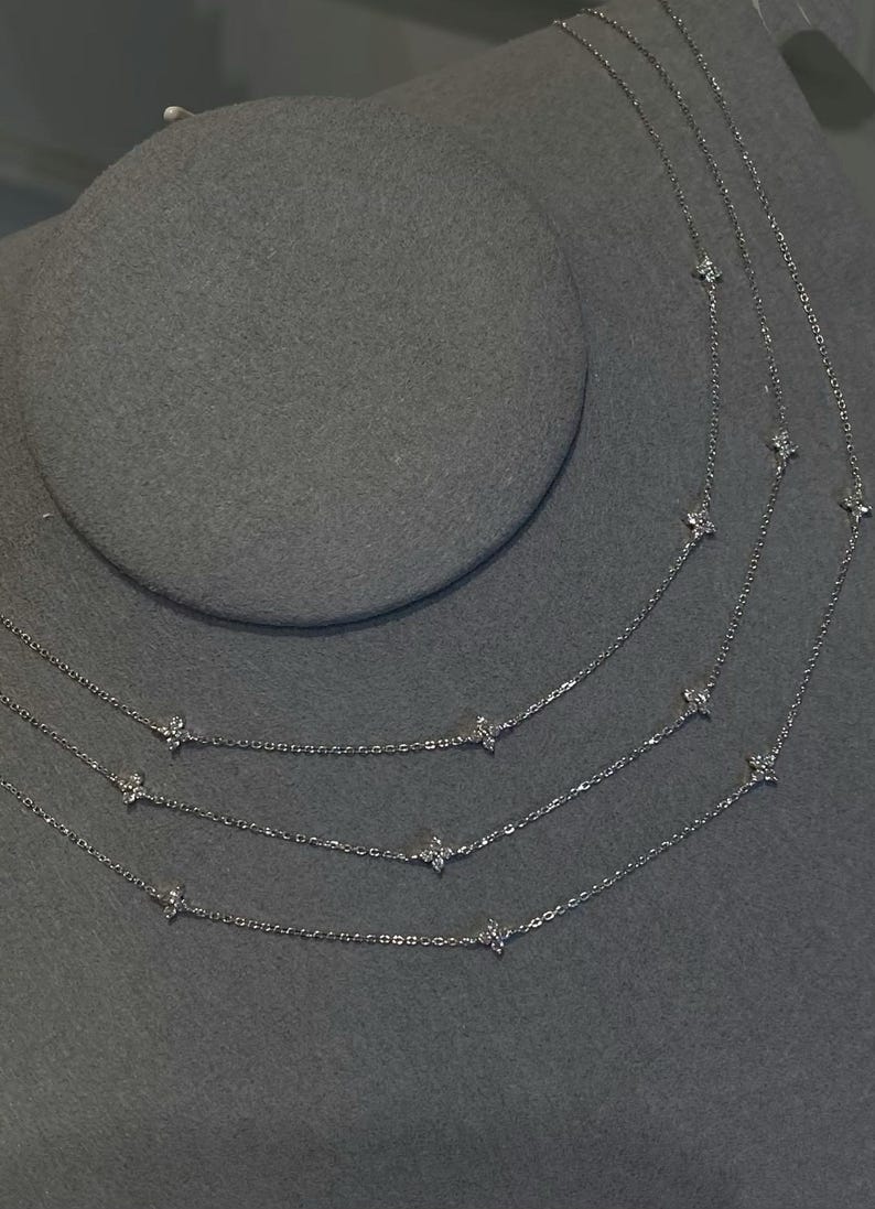 May include: Multiple delicate silver necklaces with small, sparkling flower-shaped pendants are displayed on a grey surface. The necklaces are layered, creating a tiered effect. The chains are fine and the pendants are evenly spaced.