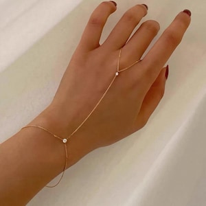 Puede incluir: Una delicada cadena de mano de oro con dos pequeñas piedras redondas y transparentes. Una piedra está situada en el centro de la sección de la pulsera y la segunda piedra se encuentra en la cadena que une la muñeca con el anillo del dedo.