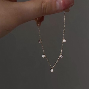 May include: Delicate gold necklace featuring small, sparkling round gemstones. The necklace has a fine chain and is held against a neutral background, showcasing its elegant design. The gemstones are evenly spaced along the chain.