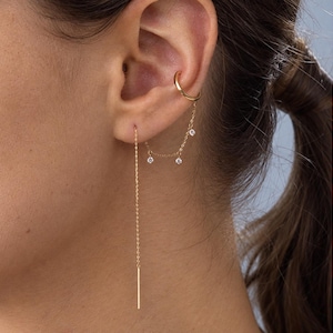 Pode incluir: Um ear cuff dourado ligado por uma corrente delicada a um brinco de fio. A corrente apresenta três pequenas pedras redondas e transparentes. O brinco de fio consiste numa corrente fina que termina numa barra dourada fina.