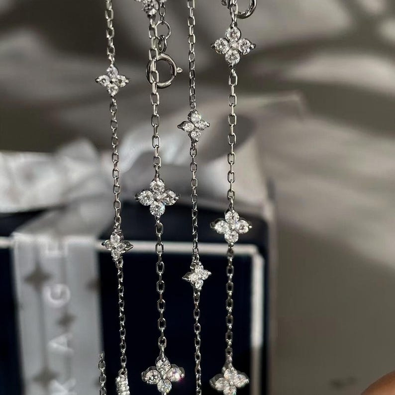 May include: A close-up shot of a silver necklace with multiple strands. Each strand is adorned with four-petal flower pendants, each set with clear, sparkling stones. The necklace is set against a blurred background.