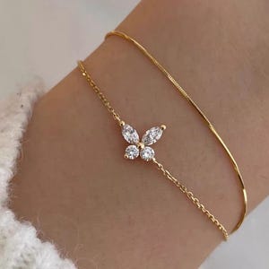 May include: A delicate gold bracelet featuring a butterfly design adorned with sparkling clear gemstones. The butterfly is the focal point, with a fine chain extending to a second, simpler gold chain. The bracelet is worn on a wrist.