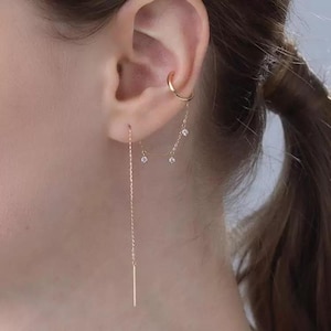 Puede incluir: Primer plano de una oreja adornada con delicadas joyas de oro. Se ve un brazalete de oreja dorado y dos pendientes. Un pendiente es una cadena larga y fina con una pequeña barra en el extremo. El otro pendiente tiene tres pequeñas gemas brillantes.