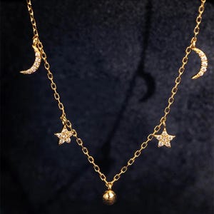 Puede incluir: Un delicado collar de oro con una cadena, un pequeño colgante esférico y colgantes. Los colgantes incluyen dos medias lunas y dos estrellas, todos adornados con piedras preciosas brillantes. El collar se muestra sobre un fondo oscuro.