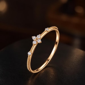 Puede incluir: Un anillo fino de oro con un motivo central de flor de cuatro pétalos engastada con pequeñas piedras claras. La banda tiene una textura granulada en el borde superior, con dos pequeñas piedras claras adicionales a cada lado de la flor.