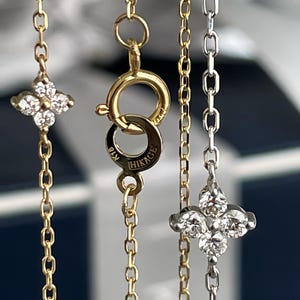 May include: Close-up of three delicate necklaces. Two are gold, one with a four-leaf clover pendant and the other with a clasp marked "HIKAGE". The third necklace is silver with a similar clover pendant. The background is blurred.
