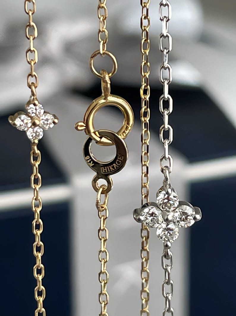 May include: Close-up of three delicate necklaces. Two are gold, one with a four-leaf clover pendant and the other with a clasp marked "HIKAGE". The third necklace is silver with a similar clover pendant. The background is blurred.