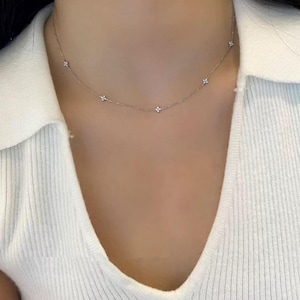 May include: A delicate silver necklace with small, flower-shaped pendants evenly spaced along the chain. The necklace is worn against a white ribbed top, creating a simple and elegant look. The necklace is a subtle accessory.