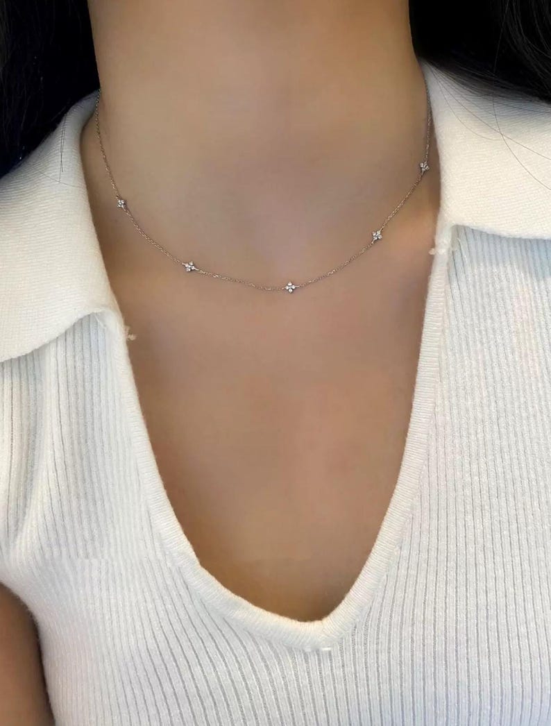May include: A delicate silver necklace with small, flower-shaped pendants evenly spaced along the chain. The necklace is worn against a white ribbed top, creating a simple and elegant look. The necklace is a subtle accessory.