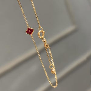 Könnte beinhalten: Eine zarte goldene Halskette mit einem kleinen vierblättrigen Kleeblatt-Anhänger mit roter Mitte. Die Kette ist fein und verstellbar, mit einem Karabinerverschluss. Die Halskette wird vor einem weichen grauen Hintergrund gezeigt.