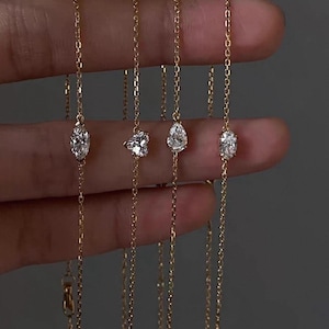 Puede incluir: Cuatro delicados collares de oro, cada uno con un colgante de piedra preciosa transparente de diferente forma. Las piedras preciosas incluyen formas ovaladas, de flor, de pera y otra forma ovalada. Los collares se sostienen sobre un fondo gris.