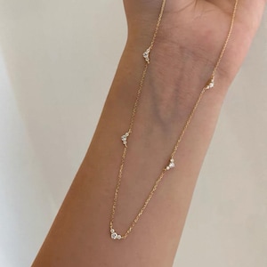 Puede incluir: Delicado collar de oro con pequeñas piedras preciosas brillantes. El collar tiene una fina cadena y está adornado con varios grupos de piedras preciosas, creando un diseño sutil y elegante. La joya se muestra en un brazo.