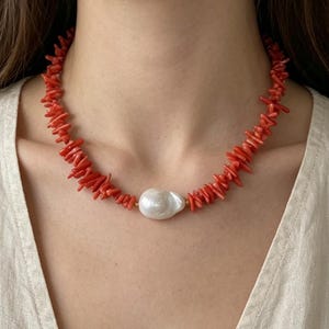 Può includere: Collana di corallo rosso con una grande perla bianca irregolare al centro. La collana è composta da piccoli pezzi di corallo rosso vivo. La perla è centrata, con dettagli dorati su entrambi i lati. La collana è indossata al collo.