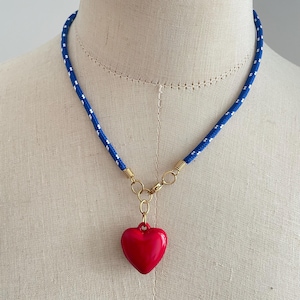 May include: A necklace featuring a blue cord with small white speckles, connected by gold-toned metal rings to a smooth, glossy red heart-shaped pendant. The cord is secured with a gold-toned metal clasp, creating a vibrant contrast between the blue, white, and red elements.
