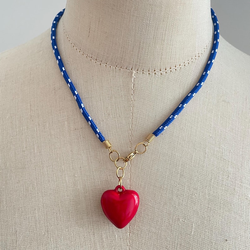 May include: A necklace featuring a blue cord with small white speckles, connected by gold-toned metal rings to a smooth, glossy red heart-shaped pendant. The cord is secured with a gold-toned metal clasp, creating a vibrant contrast between the blue, white, and red elements.