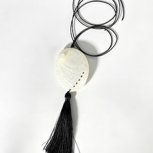Peut inclure: Un collier composé d'un grand pendentif ovale en coquille d'ormeau blanche au fini irisé naturel, percé d'une série de petits trous sur un côté. Le pendentif est suspendu à un cordon noir fin et agrémenté d'un long pompon noir assorti.