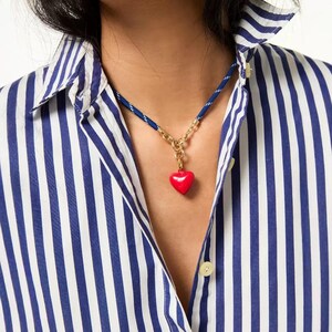 May include: A necklace featuring a vibrant red heart pendant suspended from a gold-toned metal chain link connector, which is attached to a blue and white striped cord. The cord is worn around the neck, resting against a blue and white striped shirt.