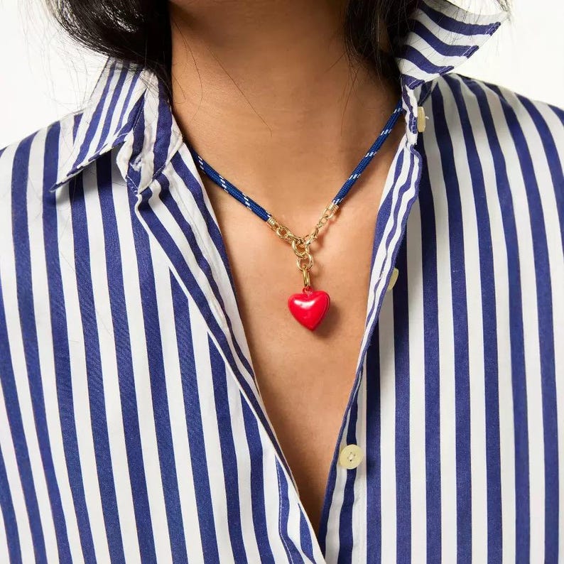 May include: A necklace featuring a vibrant red heart pendant suspended from a gold-toned metal chain link connector, which is attached to a blue and white striped cord. The cord is worn around the neck, resting against a blue and white striped shirt.