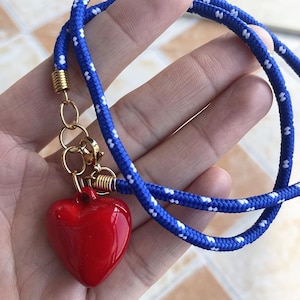May include: A necklace featuring a glossy red heart pendant attached to a blue rope cord with white flecks. The cord is secured with gold-toned metal end caps and a lobster claw clasp, connecting the pendant to the rope with gold-toned jump rings.
