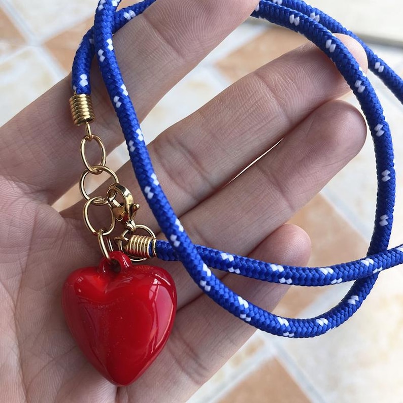 May include: A necklace featuring a glossy red heart pendant attached to a blue rope cord with white flecks. The cord is secured with gold-toned metal end caps and a lobster claw clasp, connecting the pendant to the rope with gold-toned jump rings.