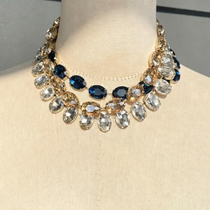 May include: A multi-layered statement necklace featuring oval-shaped crystals in shades of deep blue, champagne, and clear white. The crystals are set in gold-toned metal settings and arranged in a tiered design that rests around the neck.