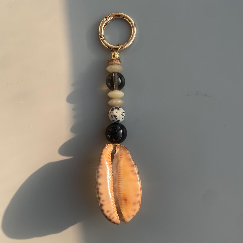 May include: A handcrafted keychain featuring a natural seashell pendant. The keychain includes a gold-colored clasp and a strand of decorative beads in various colors and textures, including black, white, and beige. The seashell is a light brown color.