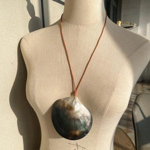 Op de afbeelding: Een ketting met een grote, iriserende schelpenhanger. De schelp is een mix van donkergroen, zwart en wit. Hij hangt aan een bruin leren koord, bovenaan geknoopt. De ketting wordt op een mannequin getoond.