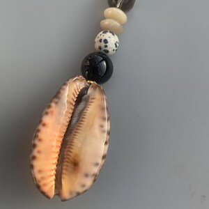 May include: A necklace featuring a large, open seashell pendant with a gradient of tan and orange hues. The necklace includes a strand of various beads, including black, white speckled, and light-colored stones.
