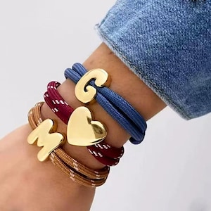 Puede incluir: Una colección de pulseras con abalorios dorados. Una pulsera presenta la letra "G", otra un corazón y una tercera la letra "M". Las pulseras están hechas de material similar a una cuerda azul, burdeos y beige.