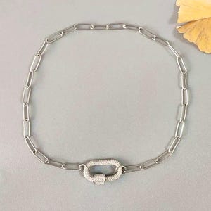 May include: Silver-toned chain necklace with an oval-shaped clasp. The clasp is encrusted with small, sparkling crystals. The necklace features a series of elongated, rectangular links. The background is a solid, light gray color.