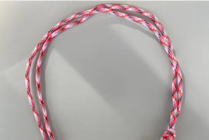 May include: A length of braided cord featuring a multi-colored pattern of pink, white, black, and grey threads. The cord is arranged in a loose loop, showcasing the intricate woven texture and the vibrant combination of these four distinct colors.