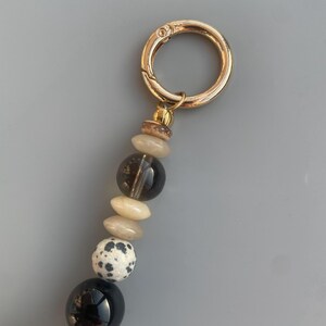 May include: A gold-colored metal clasp attached to a beaded charm. The charm features a variety of beads, including black, speckled white, and light-colored stones. The background is a solid gray.