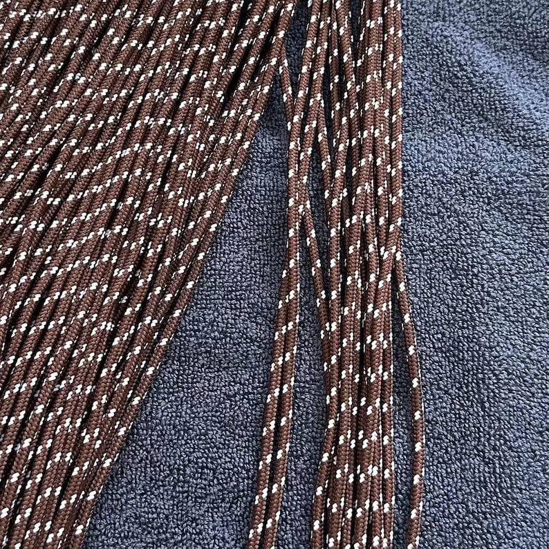 May include: A collection of brown paracord strands featuring a repeating white diagonal fleck pattern. The cords are arranged in parallel lines, showcasing the braided texture and the consistent color scheme of the individual rope segments.