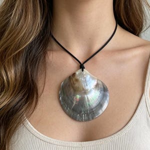 May include: A large, iridescent shell pendant necklace. The shell is a mix of gray, brown, and pearlescent colors, suspended from a black cord. The necklace is worn against a neutral-toned top.