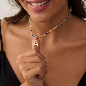 May include: A vibrant beaded necklace with a gold-coloured letter "A" pendant. The necklace features small, multi-coloured beads in various hues. The pendant is suspended from a clear, round bead. The necklace is worn around the neck.