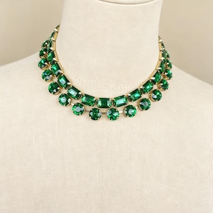 May include: A double-strand necklace featuring faceted emerald green crystals set in gold-toned metal. The design consists of two rows of alternating rectangular and round stones, creating a layered appearance around the neck of the display form.
