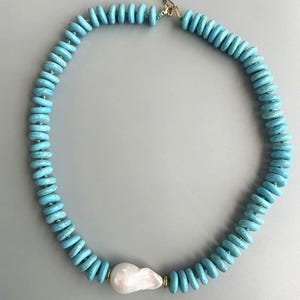 May include: A turquoise beaded necklace with a large, irregular white pearl pendant. The necklace features round, flat turquoise beads strung together with a gold clasp. The pearl is off-white and has a unique, organic shape.