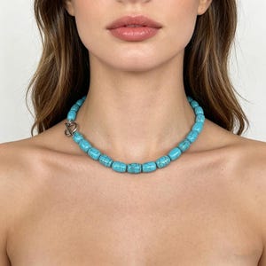 May include: A turquoise beaded necklace with a silver clasp. The necklace features oval-shaped beads in a vibrant blue-green hue. The clasp is a silver toggle closure. The necklace is worn around the neck.