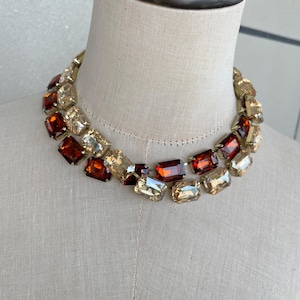 May include: A multi-strand statement necklace featuring rectangular faceted crystals in alternating shades of deep amber and champagne gold. The stones are set in a gold-tone metal frame, creating a layered collar design that sits around the neck.