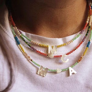 Può includere: Tre collane a strati con ciondoli a forma di lettera. La collana superiore è rossa, gialla e bianca, quella centrale è viola e rossa, e quella inferiore è verde e gialla. Ogni collana ha un ciondolo a forma di lettera e un ciondolo a cuore.