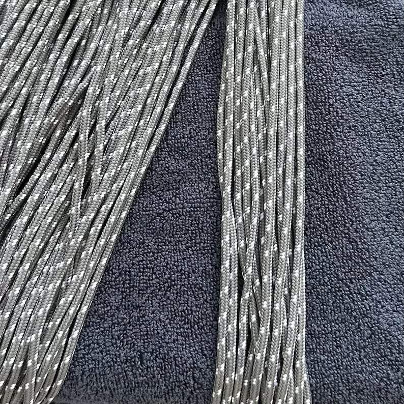 May include: A collection of multiple strands of grey cord featuring a repeating white speckled pattern. The cords are arranged in parallel vertical lines, showcasing a braided texture and a consistent diameter throughout each individual length.