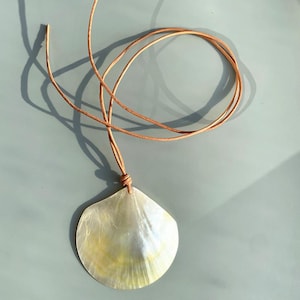 Op de afbeelding: Een parelmoeren schelpvormige hangerketting met een bruin leren koord. De hanger heeft een natuurlijke, iriserende glans met gele en witte tinten. Het koord is aan de basis van de hanger geknoopt, ongeveer 50 cm lang.